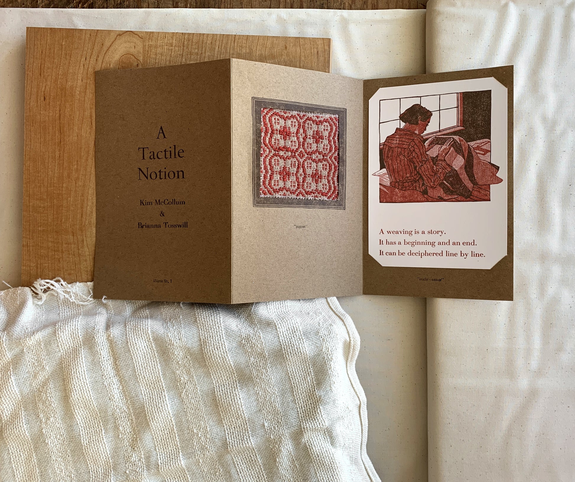 A tri-fold booklet lies on a layered white linen surface. The booklet says "A Tactile Notion / Kim McCollum & Brianna Tosswill / Vol. 1" on the front. The second page has a window with a red and light brown weaving sample which invites touch. The third page has a linocut print of a woman looking intently "reading" the weave of the blaket on her lap. Everything is in tones of brown, pink, red, and beige.