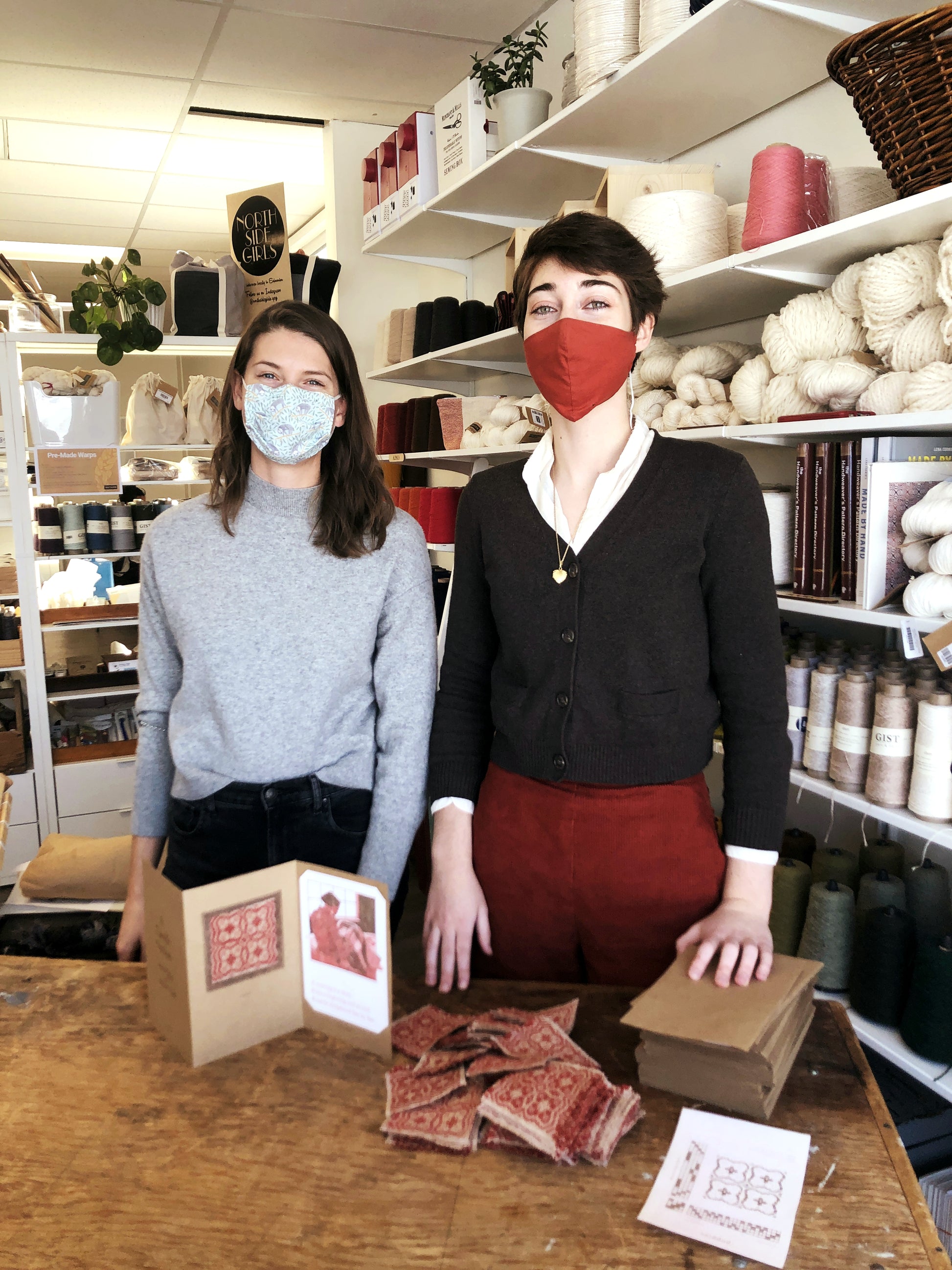 A photo of Kim McCollum and Brianna Tosswill in a weaving studio with A Tactile Notion on the table in front of them. They are both tallish, white, brunette women (no relation). They are both wearing fabric masks because this photo was taken in 2020.